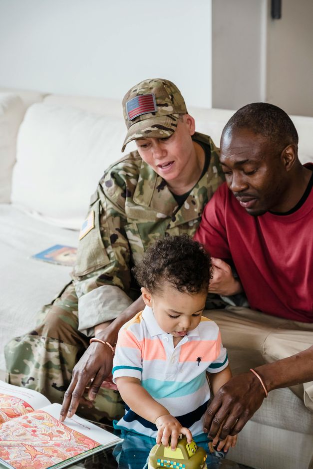 Veteran Couple and a toddler playing together on a couch, with toys and a wrapped gift nearby.