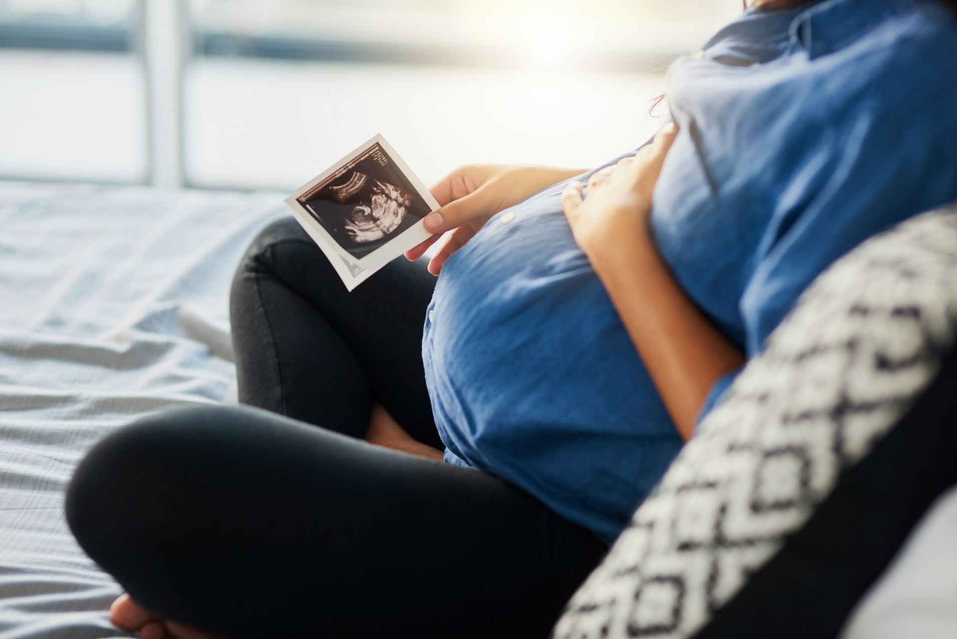A pregnant woman is sitting on a bed holding an ultrasound picture of her baby.