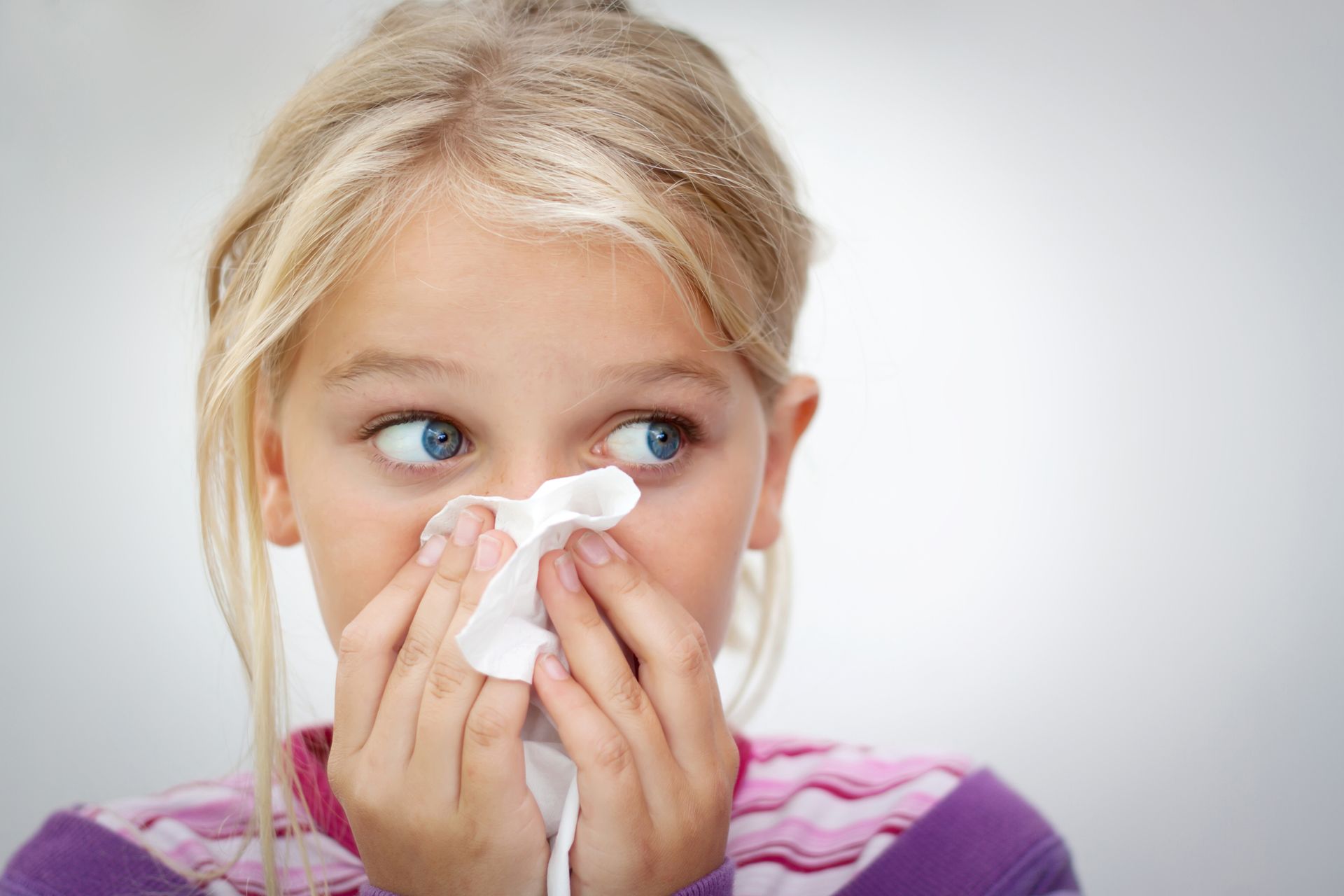 Girl with blonde hair blowing her nose with a tissue. She looks to the side.
