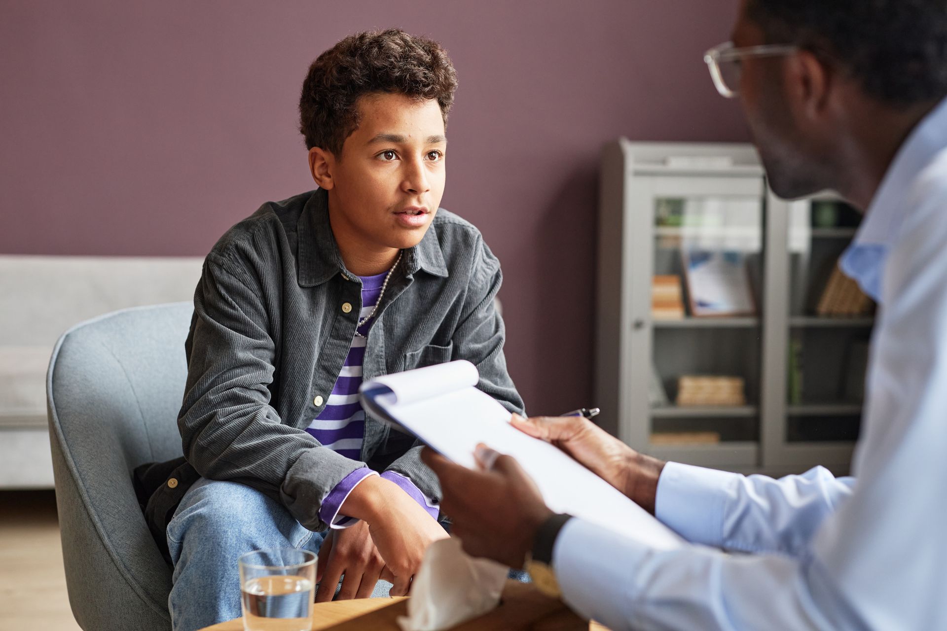 Teenager in therapy session with therapist, talking, holding a notepad.