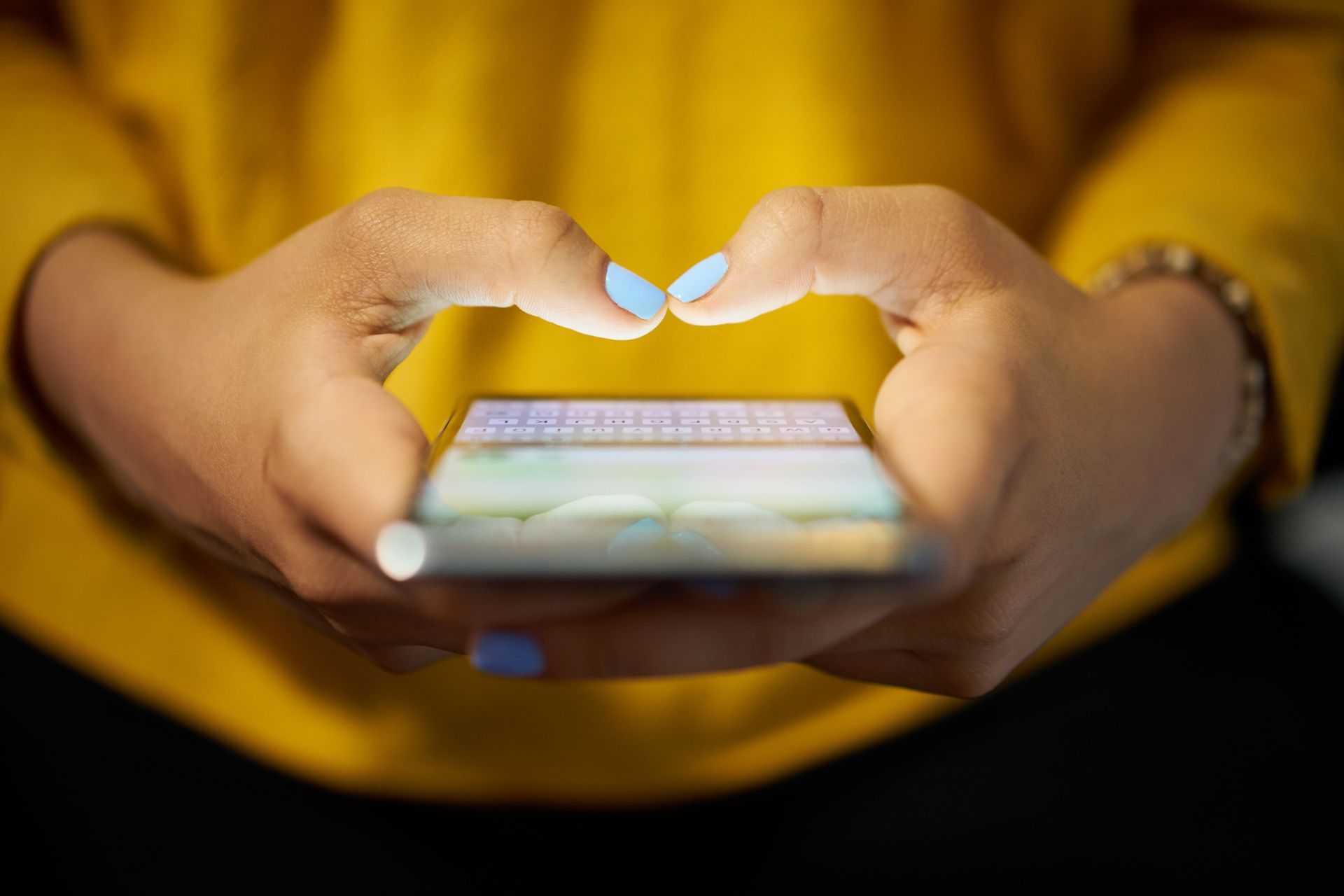 Hands holding a smartphone with blue nails, screen illuminated, against a yellow shirt.