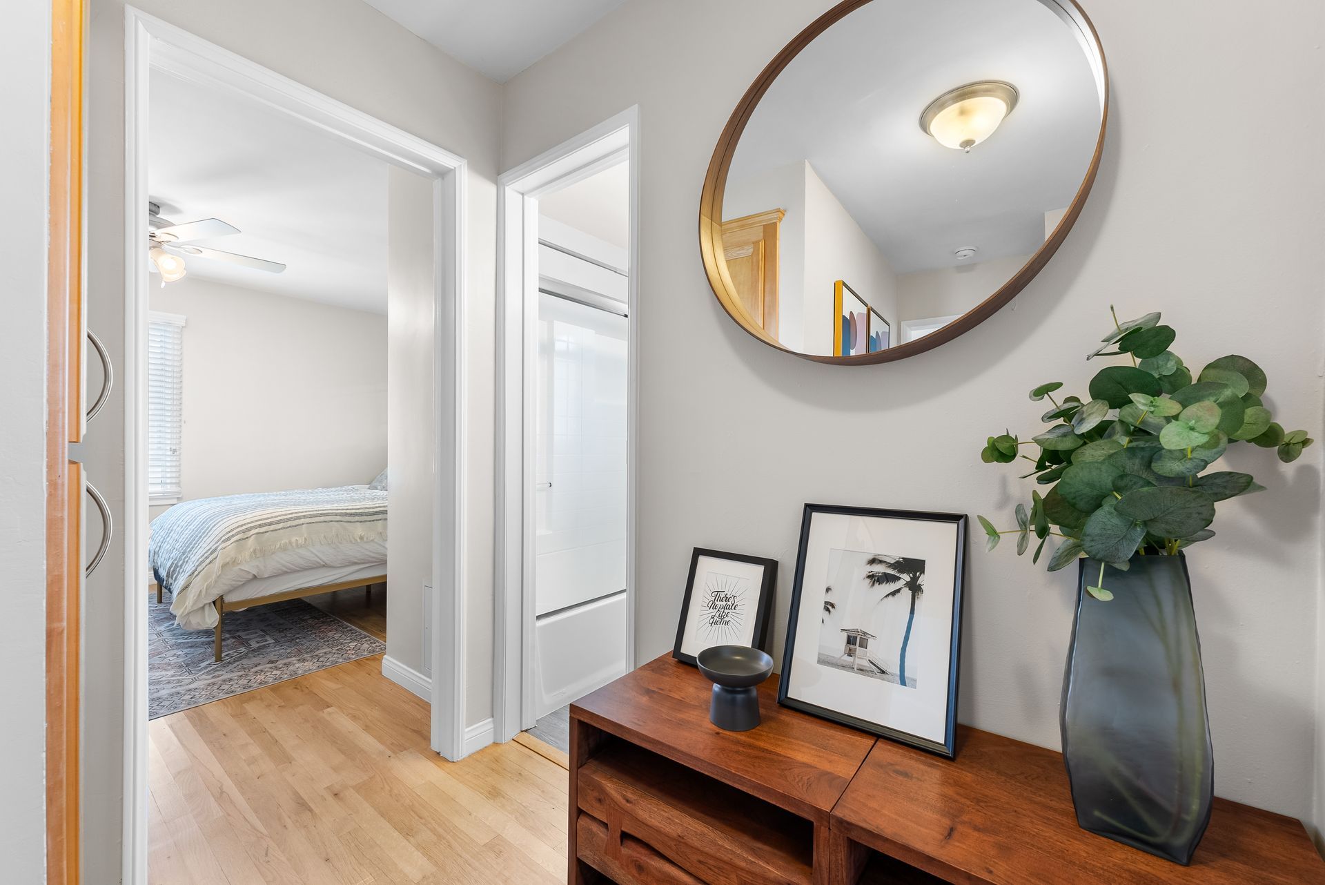 Hallway with wood floor, console table with art, round mirror, and leafy vase.