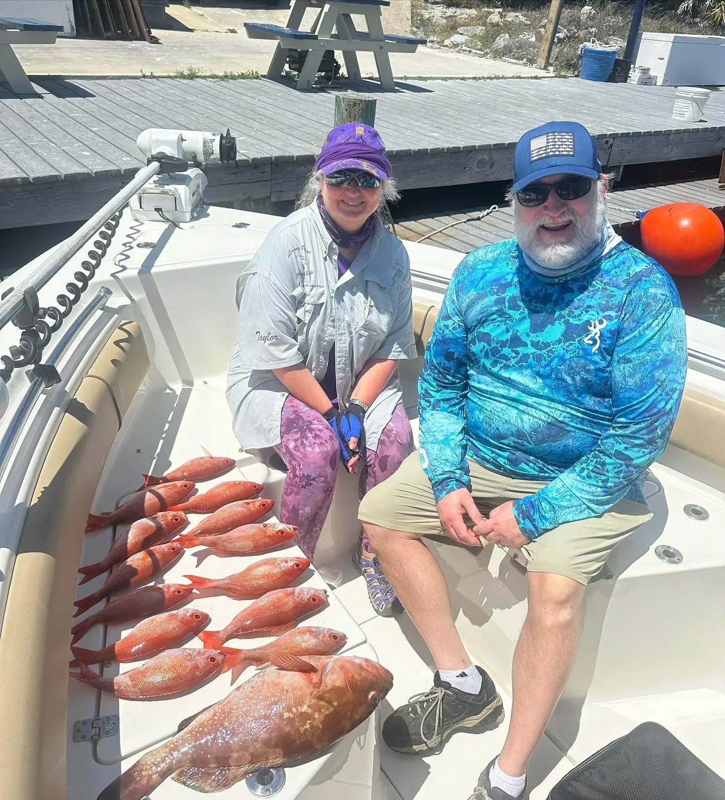 Two people sit on a boat deck beside a line of red fish, smiling near a dock.