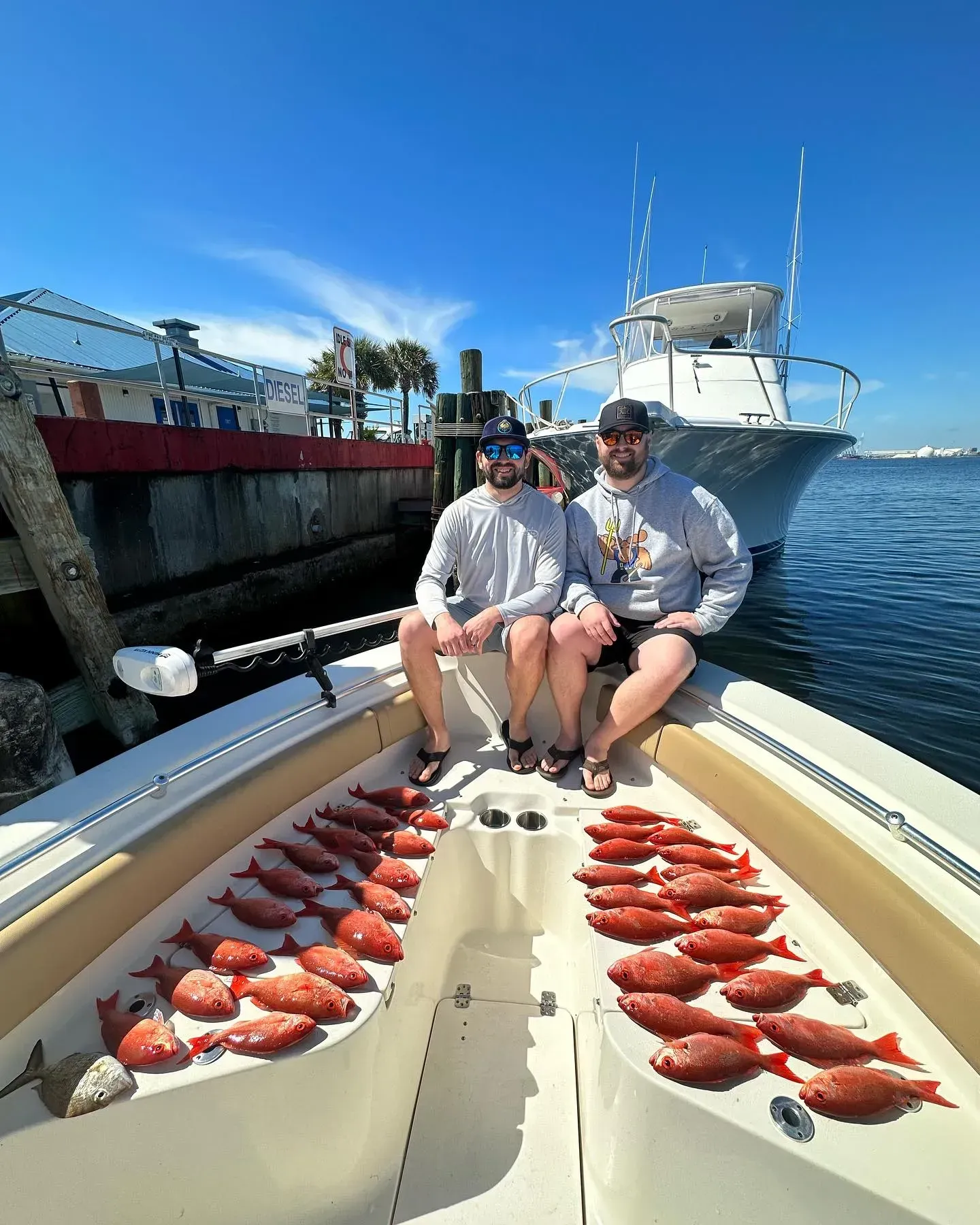 Two people on a boat with rows of red fish on deck beside a docked yacht under a blue sky