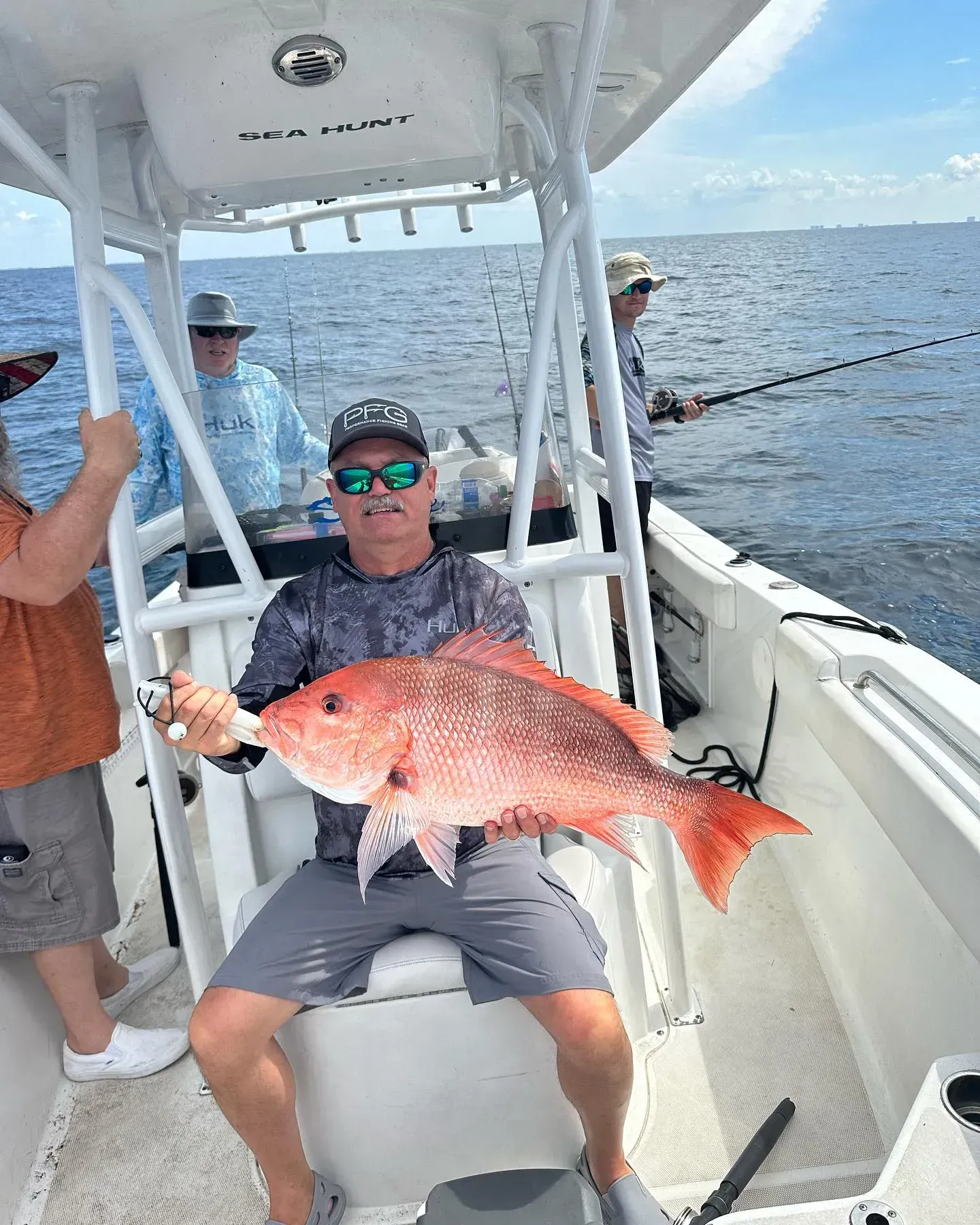 Man on a boat holding a large red fish, with two anglers and open water behind him.