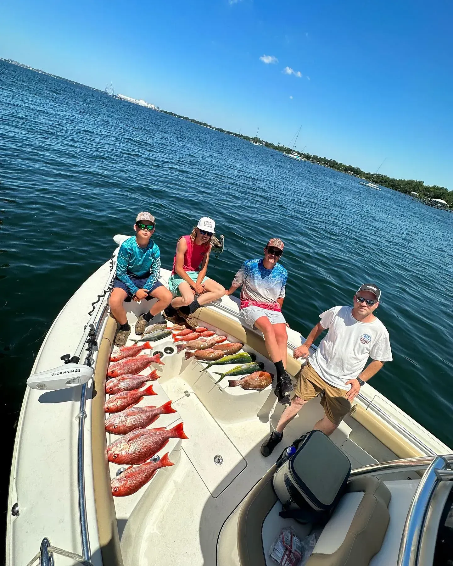 Four people on a fishing boat with a catch of red fish on deck, on open water under a blue sky.