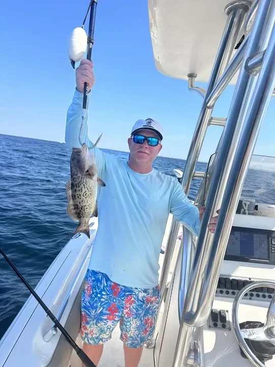 Person on a boat holding up a large fish over the ocean, wearing sunglasses and a white shirt.
