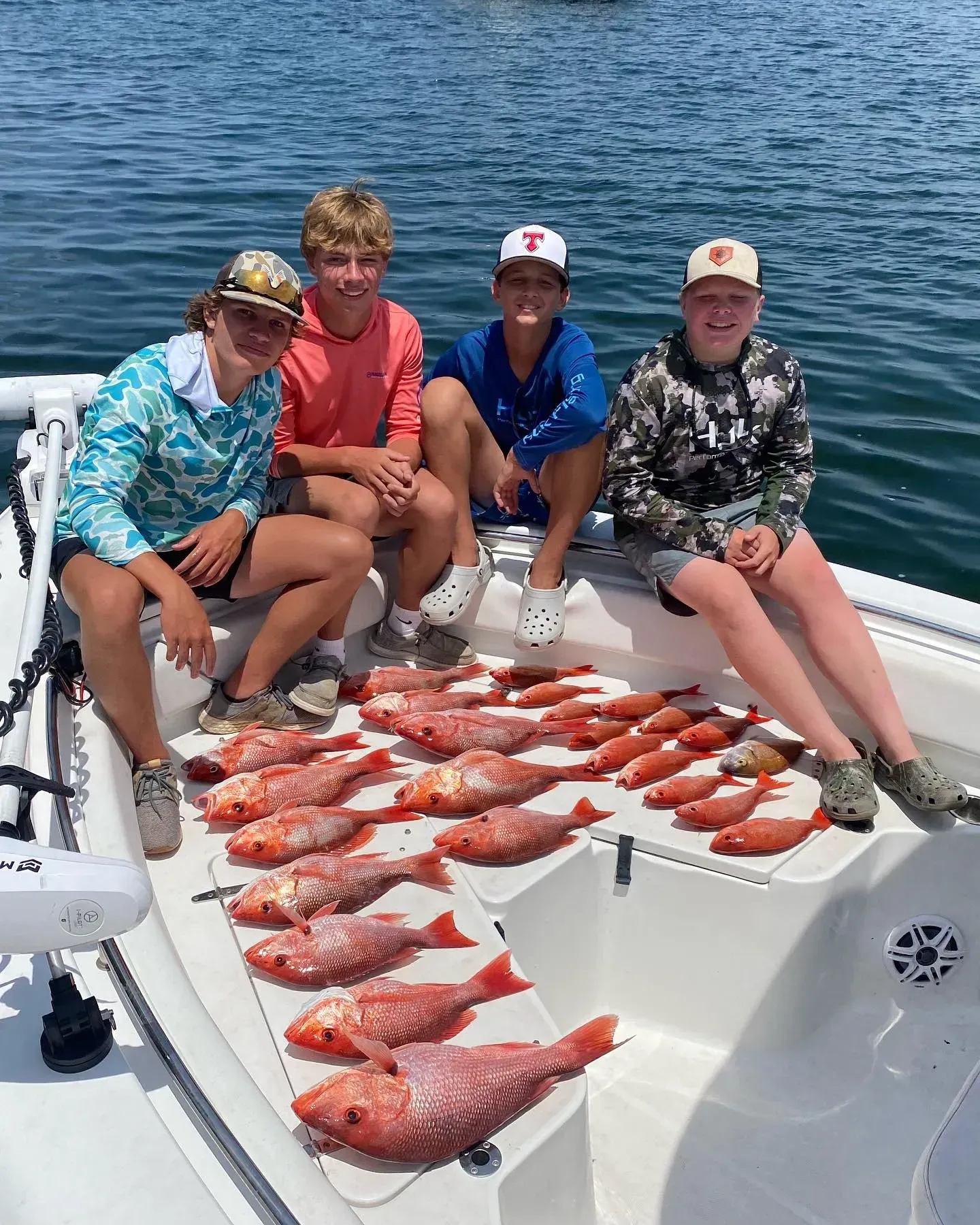 Four people sit on a boat with a large catch of red fish spread on deck, water in the background.