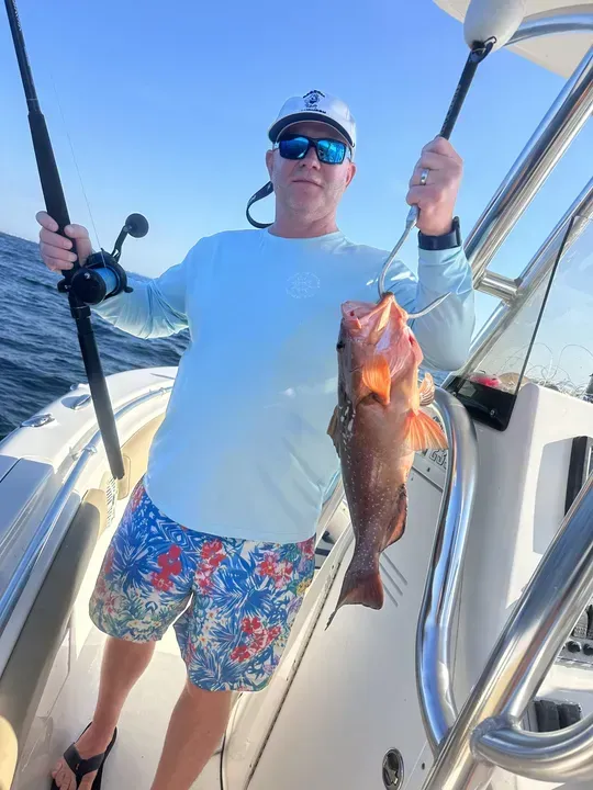 Man on a boat holding fishing rods and a large fish, wearing a sun shirt and colorful shorts.