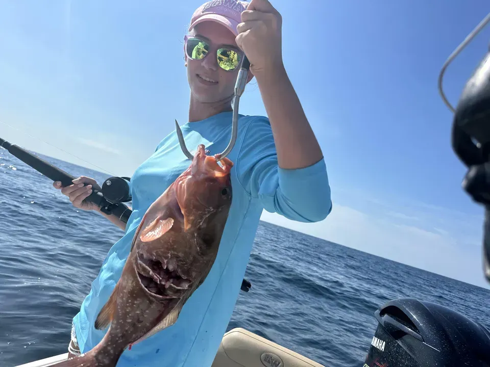 Person holding a large fish on a boat at sea, wearing a blue shirt and reflective sunglasses.