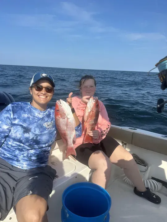 Two people on a boat hold up large red fish, with the ocean in the background.