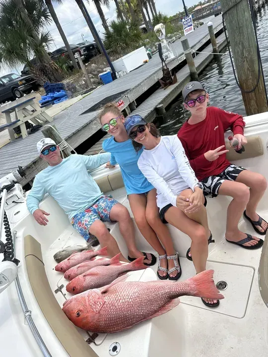 Four people sit on a dock beside several large red fish, smiling in sunglasses near the water.