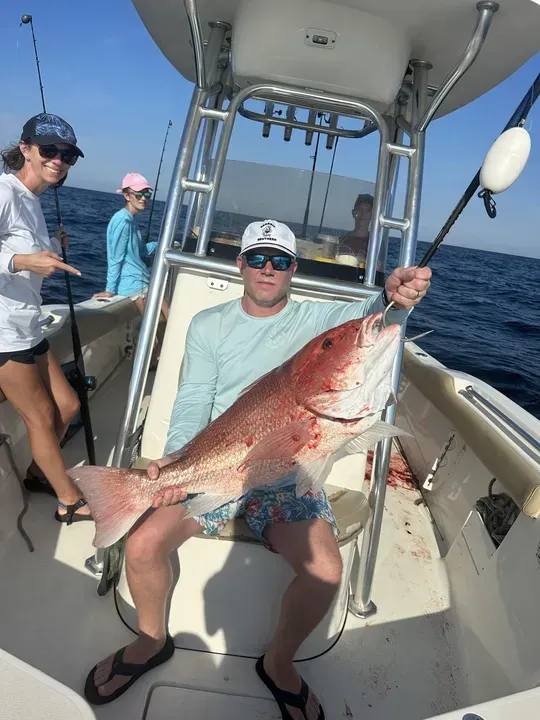 Man on a boat holding a large red fish, with two others nearby on the open ocean.