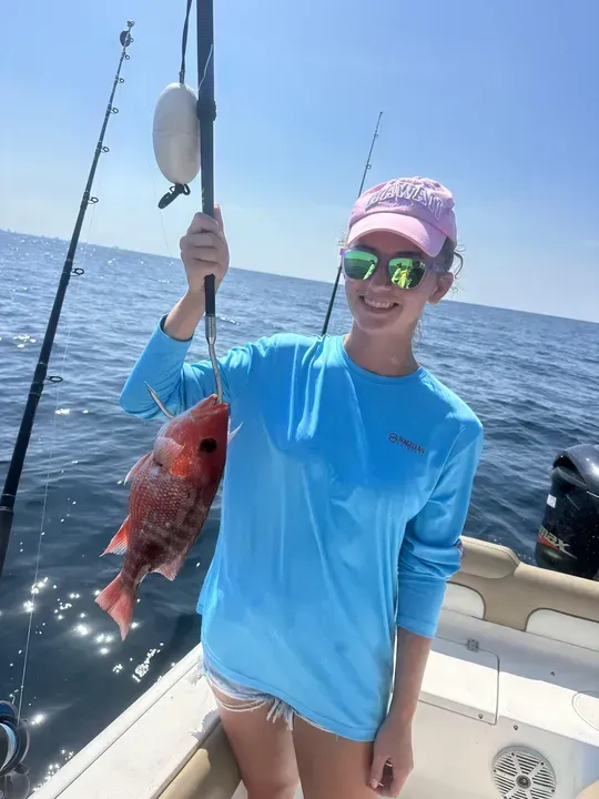 Person on a boat holding a red fish on a fishing line, wearing a blue shirt and cap on the ocean.