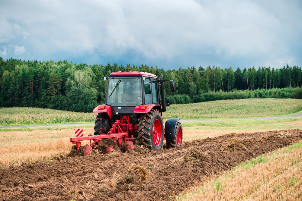A Red Tractor is Plowing a Field on a Cloudy Day — SMH Valley Fencing & Maintenance In Pokolbin, NSW