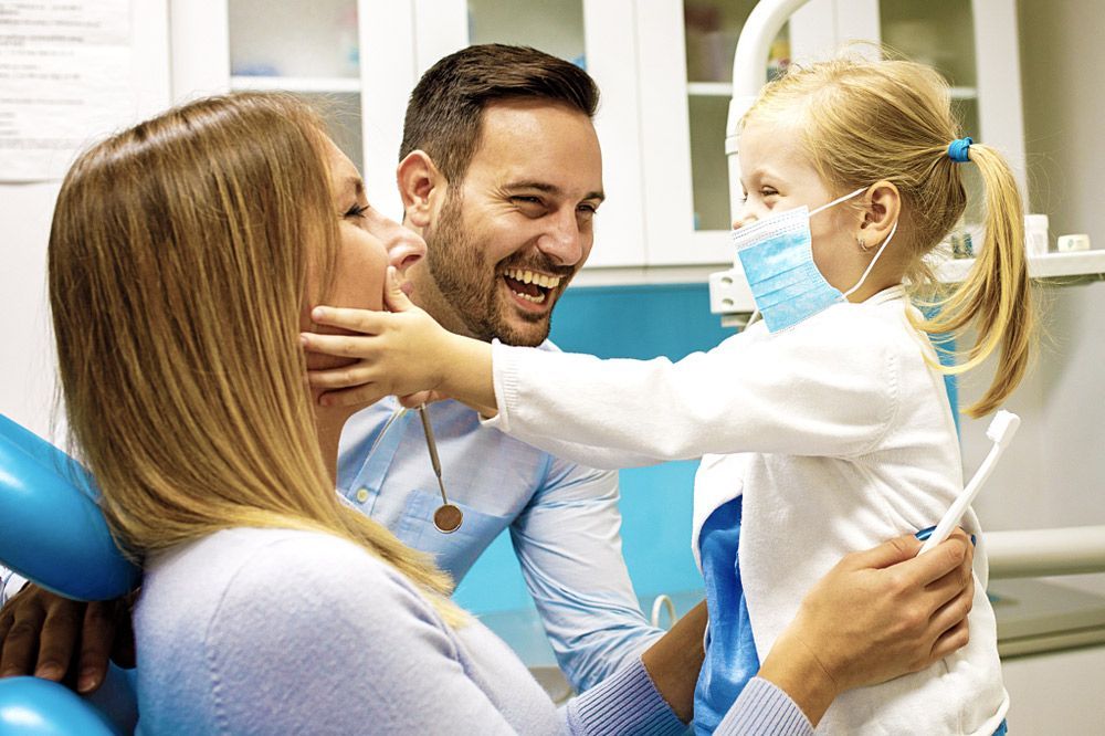 A Little Girl Is Holding A Toothbrush While Sitting In A Dental Chair With Her Parents — Regal's Dental In Yeppoon, QLD