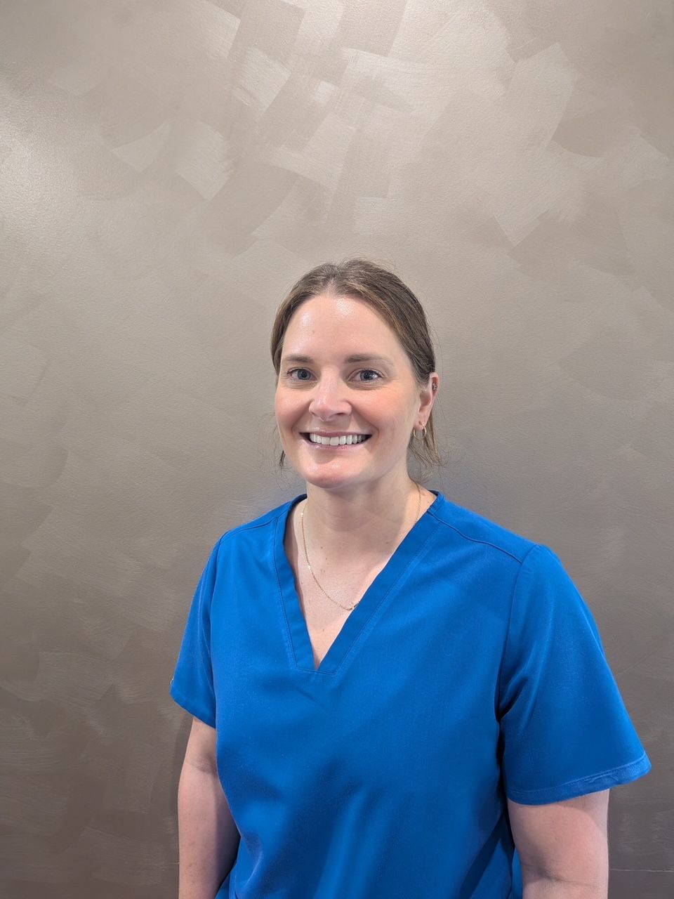 Person in a blue scrub top smiling against a neutral textured background — Regal's Dental In Yeppoon, QLD