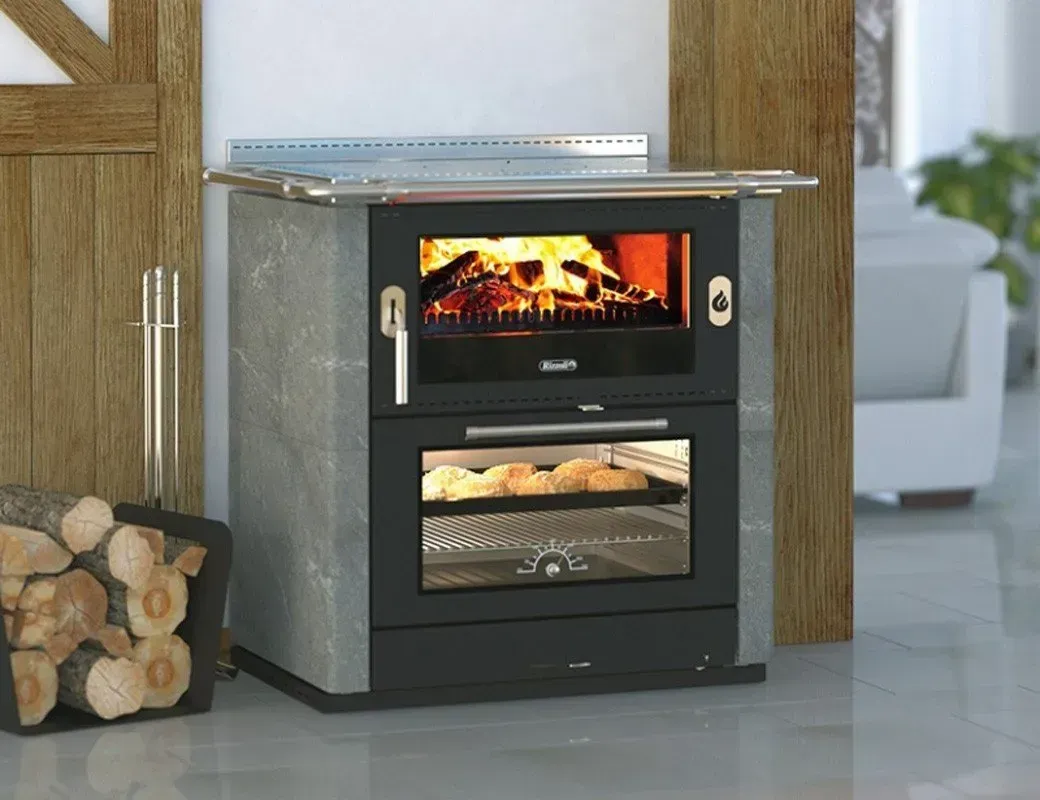 Stufa a legna compatta di colore nero con fiamme visibili, posizionata all'interno accanto a una catasta di legna.
