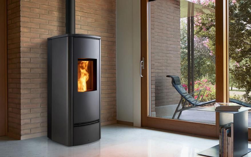 Stufa a legna moderna di colore nero con fiamma visibile, in una stanza luminosa accanto a grandi finestre.