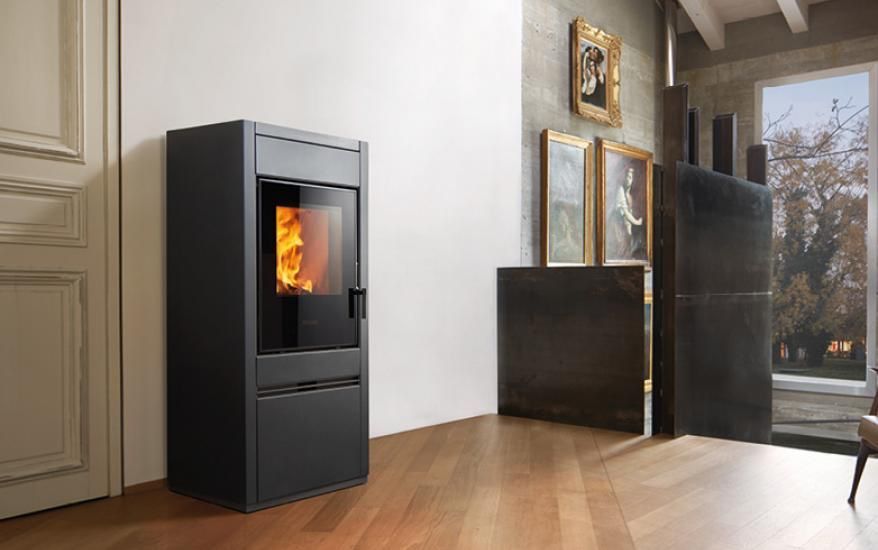Una moderna stufa a pellet nera illumina una stanza luminosa con pavimento in legno e ampie finestre.