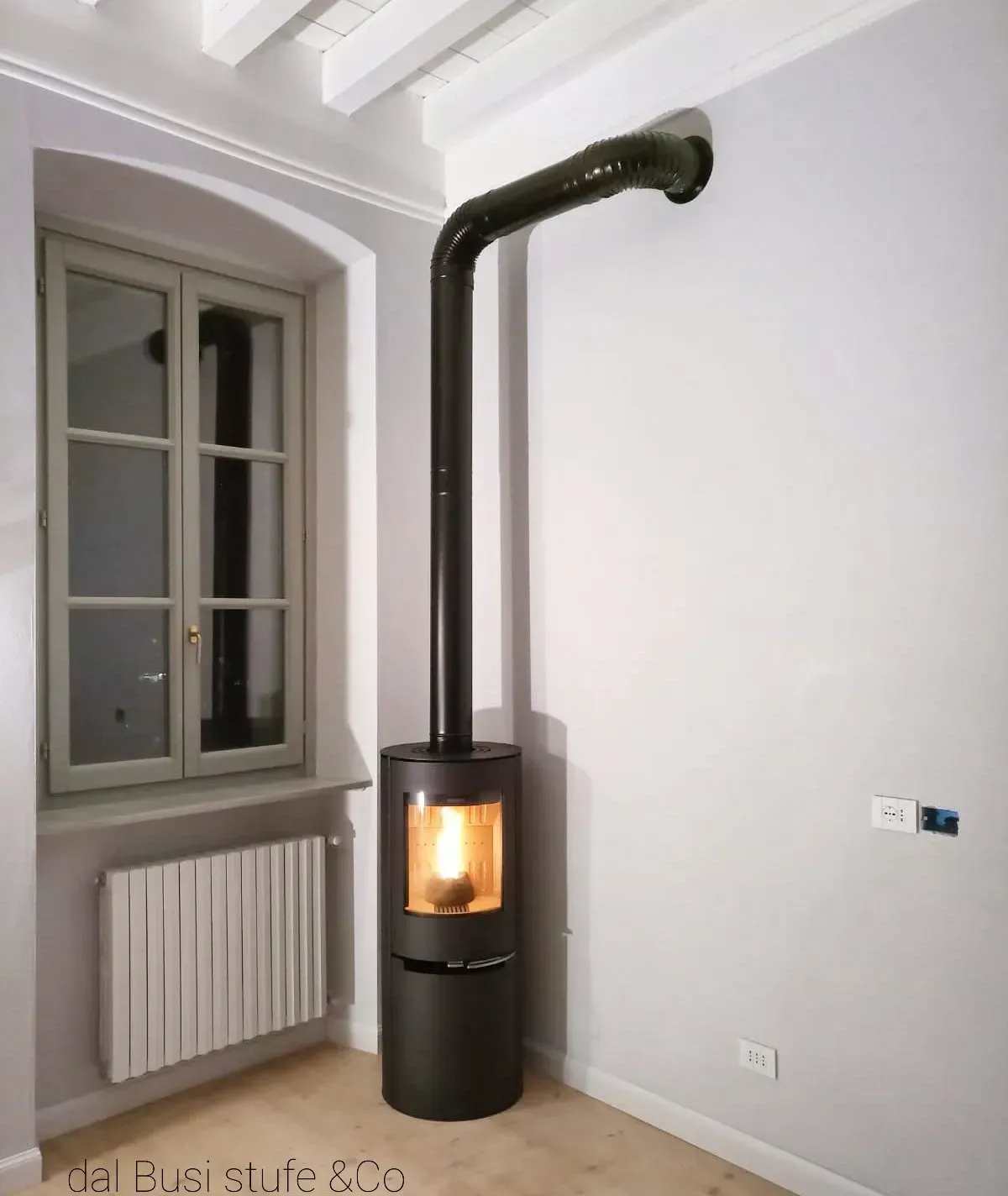 Stufa a legna nera con canna fumaria alta in una stanza bianca accanto a una finestra e un termosifone.