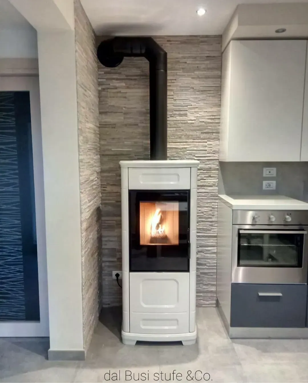Stufa a pellet bianca con fuoco acceso in una cucina moderna, accanto a un forno grigio e piastrelle a parete.
