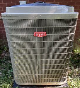 Outdoor Bryant central air conditioner unit beside a brick wall, light gray with red logo.
