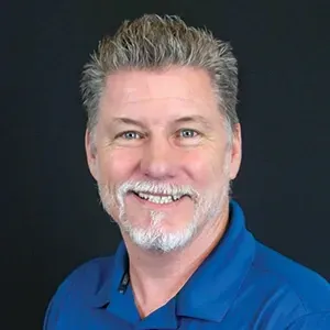 Smiling man with gray hair and beard in a blue shirt against a dark background