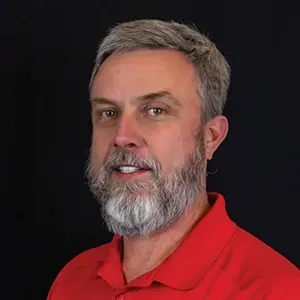 Portrait of a gray-haired man with a beard wearing a red polo shirt against a black background