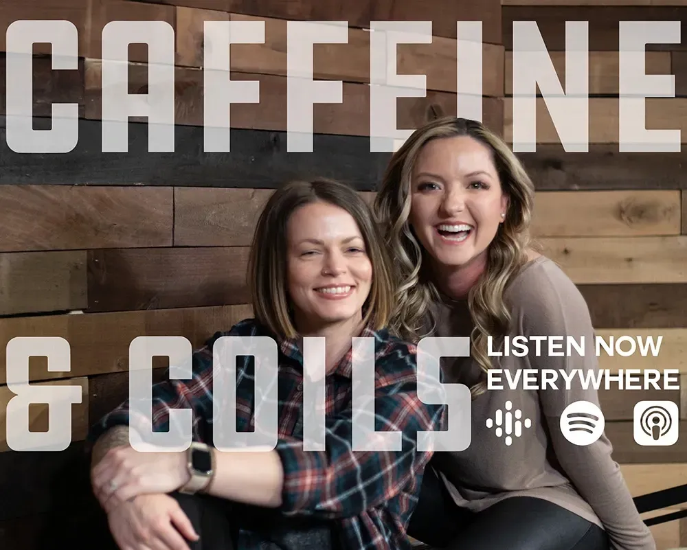 Two smiling people posing in front of a wood wall with “CAFFEINE & COILS” and “LISTEN NOW EVERYWHERE” text.
