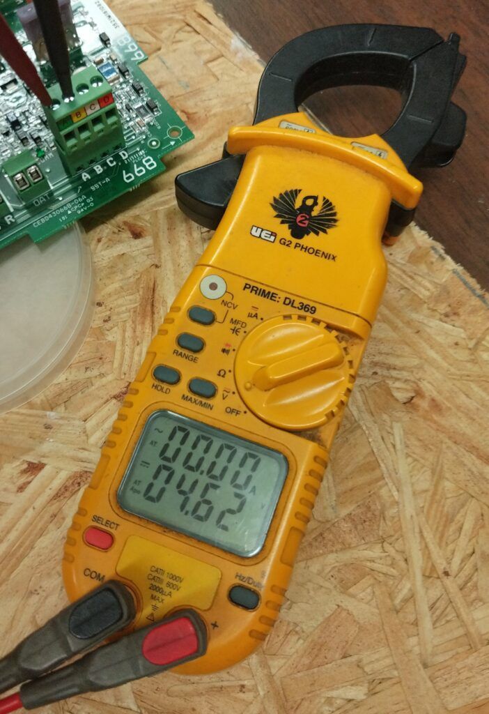 Yellow clamp meter on a wooden surface next to circuit boards, showing a digital reading.