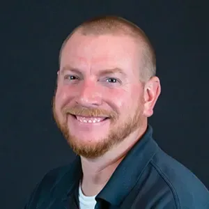 Smiling person with short red hair and beard on a dark background, wearing a dark jacket