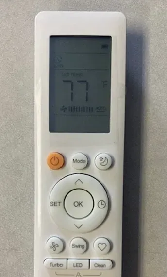 White air conditioner remote with LCD showing 77°F and orange power button on a light background