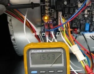 Digital multimeter reading 15.53 in an electrical panel with multicolored wires and a lit circuit board LED.