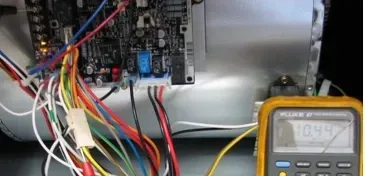 Wiring connected to a circuit board inside an electronics enclosure, with a multimeter in the foreground