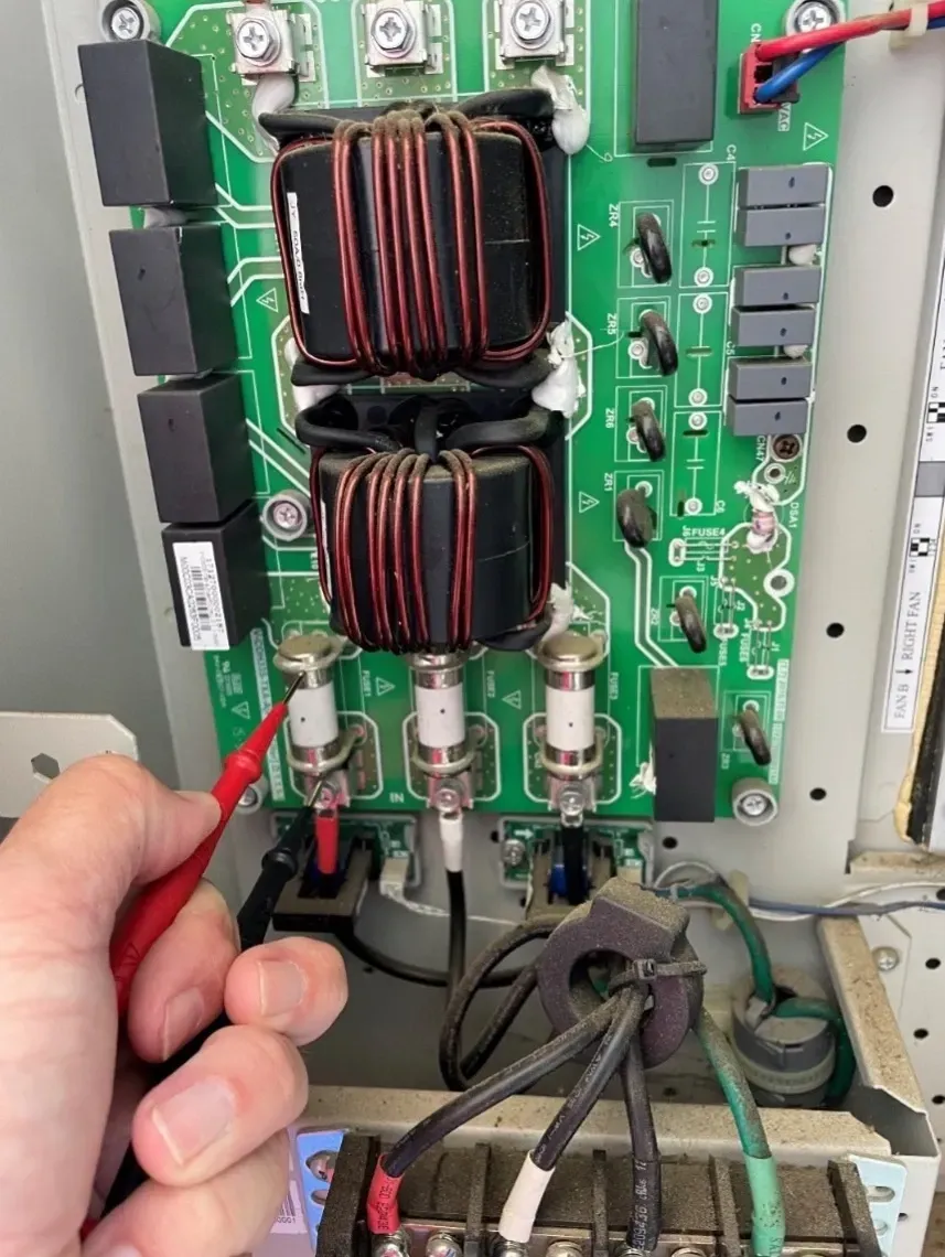 Hand holding test probes near a green circuit board with copper coils and wires inside electronics equipment