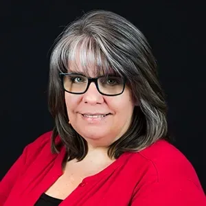 Portrait of a smiling person with gray-streaked hair, black glasses, and a red top against a black background