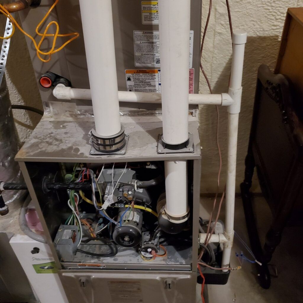 Open furnace cabinet with exposed wiring, pipes, and blower components in a utility room