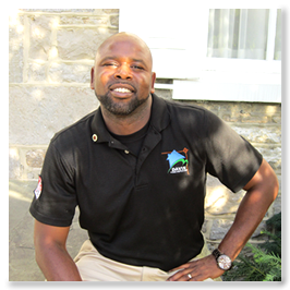 Man in black polo shirt with logo, smiling, sitting, leaning against a stone wall.