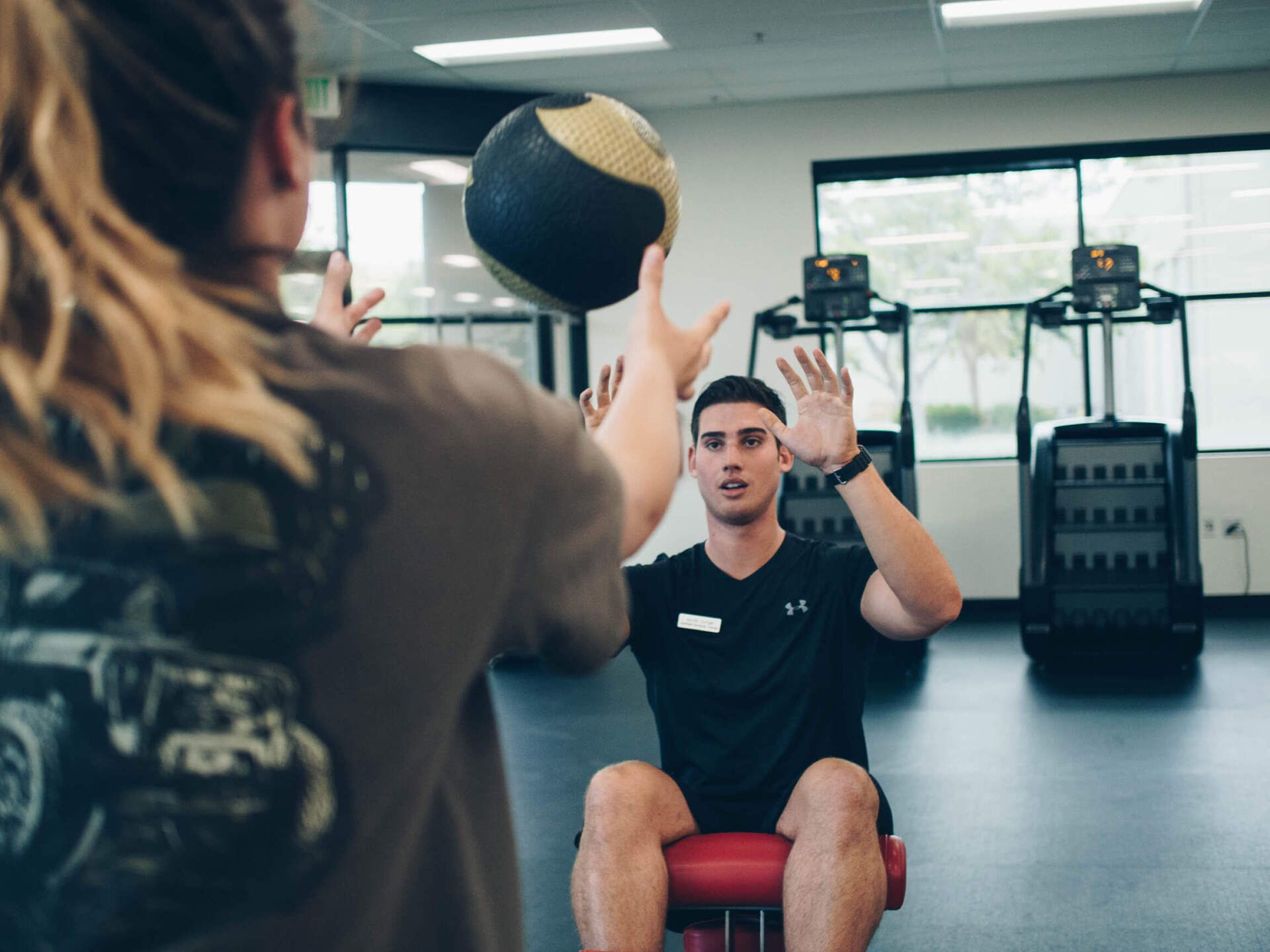 A man is sitting on a chair in a gym while a woman throws a medicine ball to him.