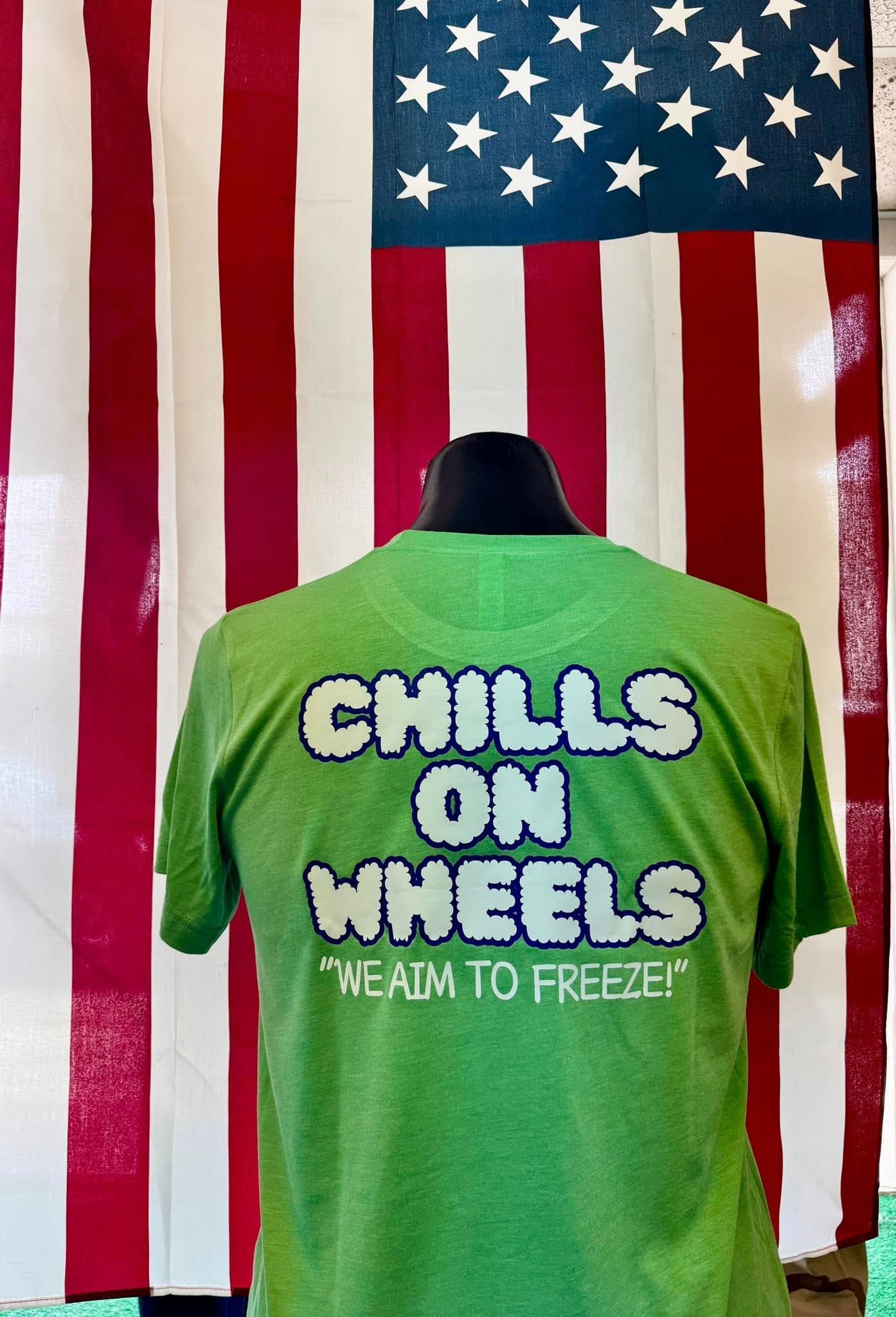 A green t-shirt that says chills on wheels is on a mannequin in front of an american flag.