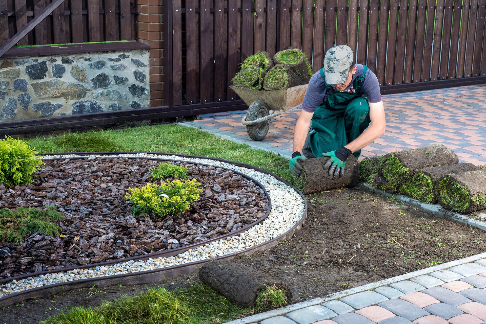 Person planting soil in a landscaped garden bed beside a paved walkway and wooden fence