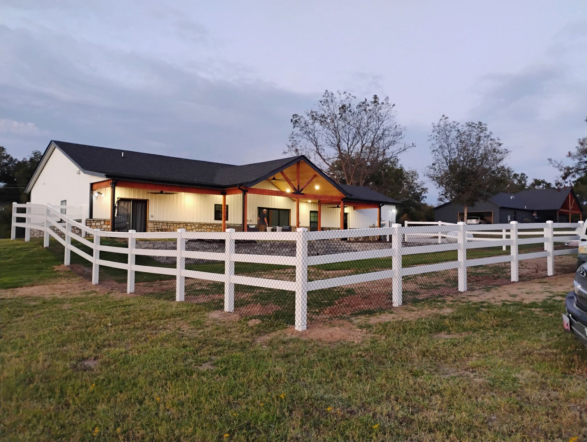 White fenced ranch house at dusk with warm porch lights and a cloudy sky