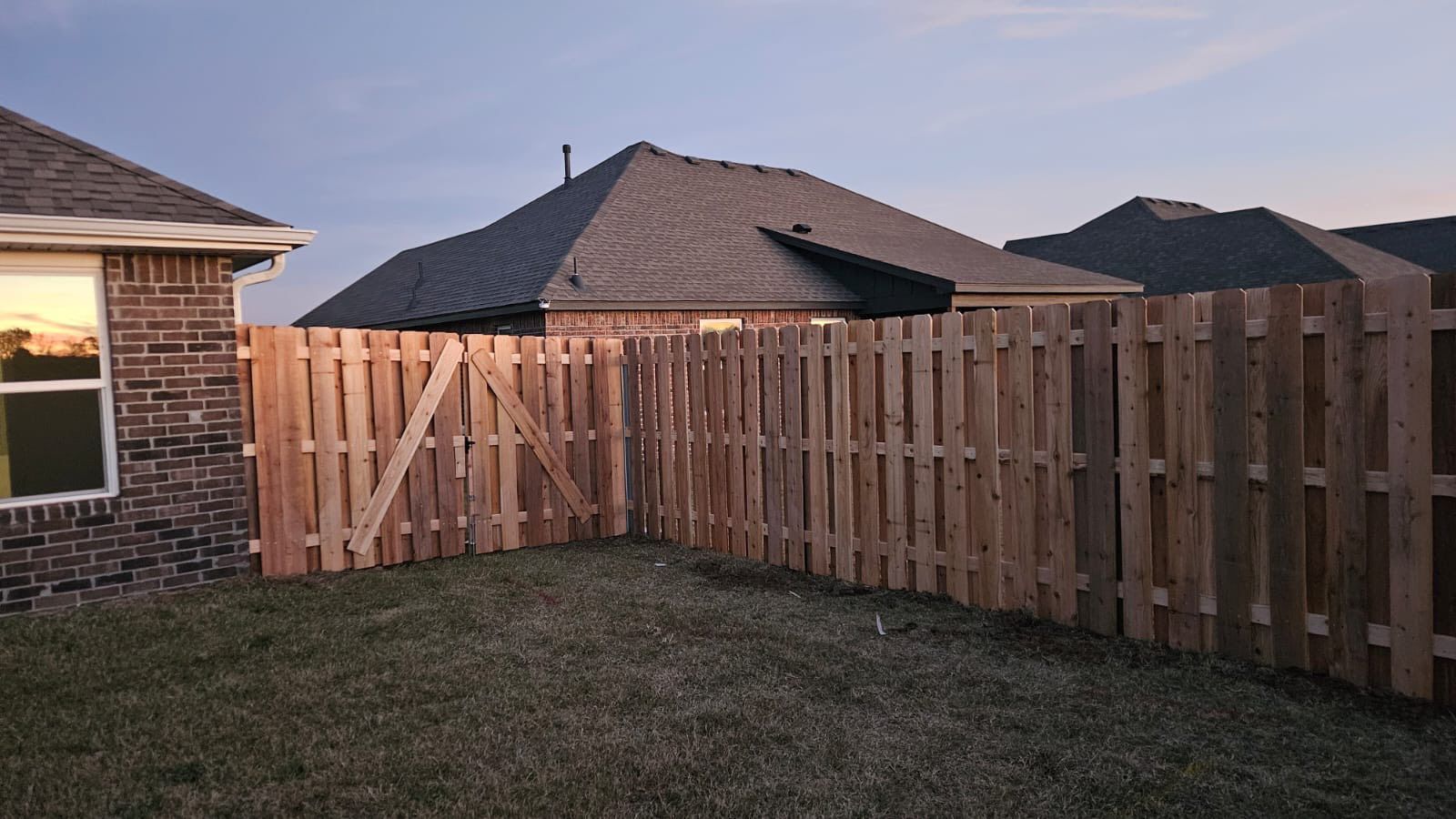 Wooden privacy fence enclosing a backyard beside a brick house at dusk.