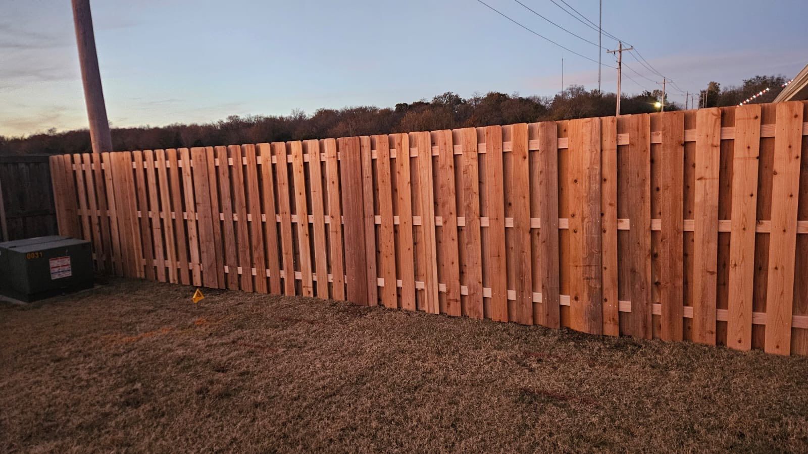 Wooden privacy fence along a grassy yard at sunset, with utility poles and wires behind it.