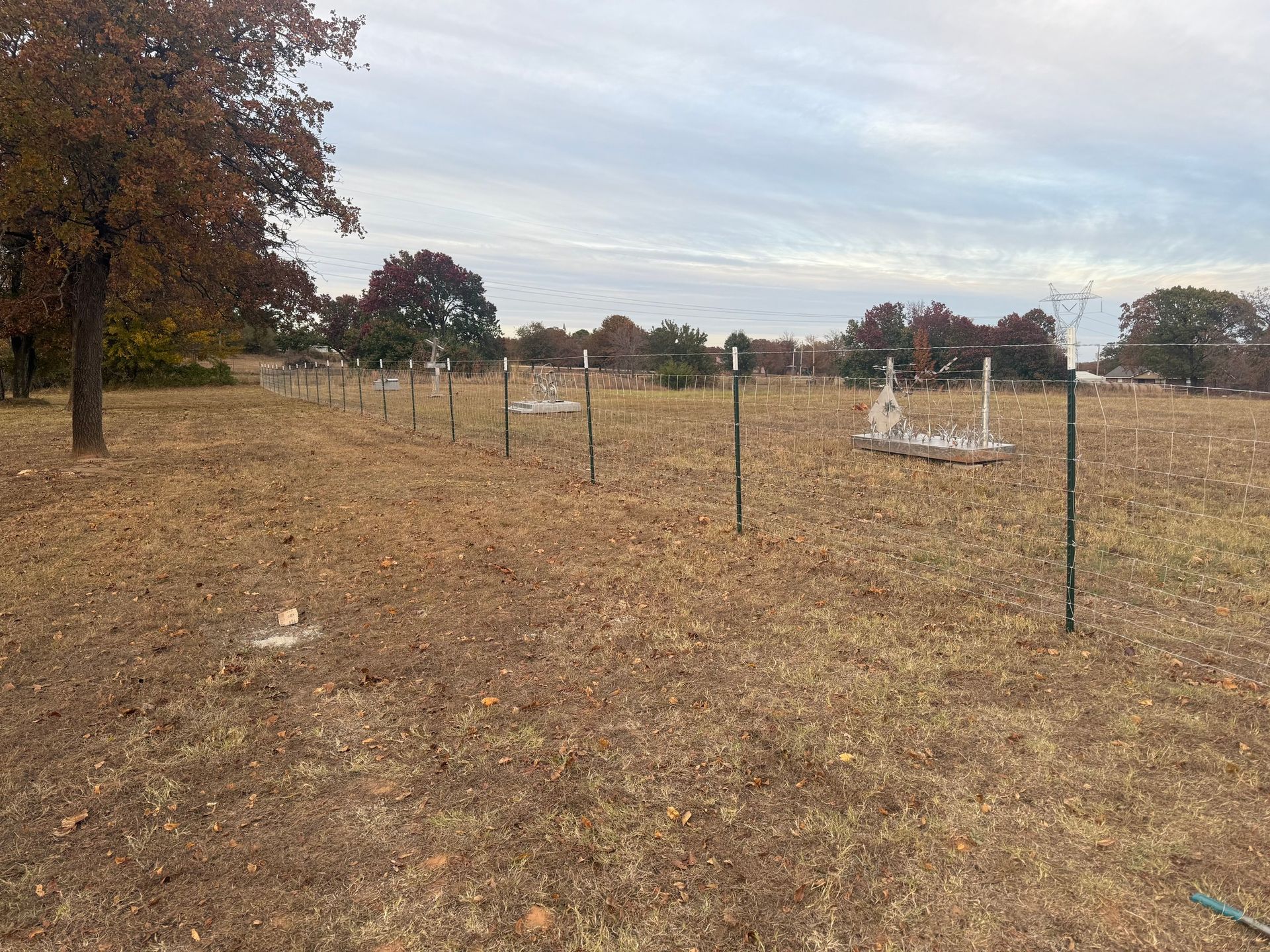 Rural field with dry grass, scattered fence posts, and a few white plastic-covered structures under a cloudy sky