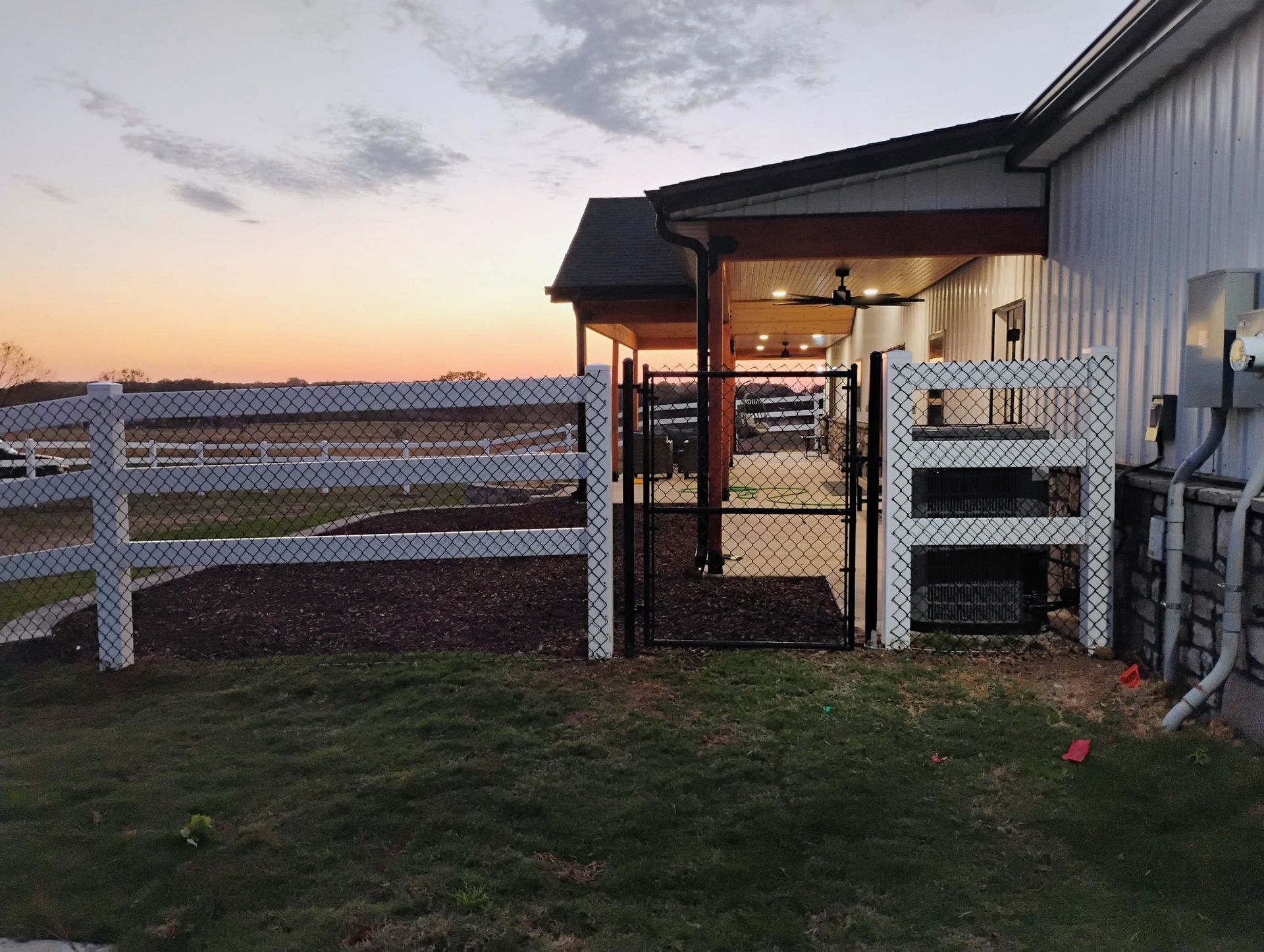 Backyard at sunset with white fence, open gate, and house porch lit warmly