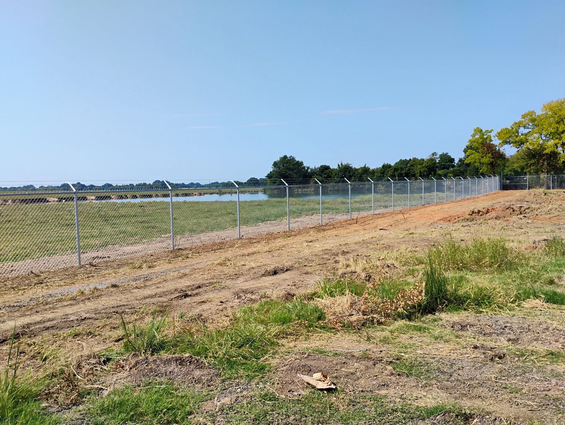 Rural dirt road beside a wire fence and open field under a clear blue sky