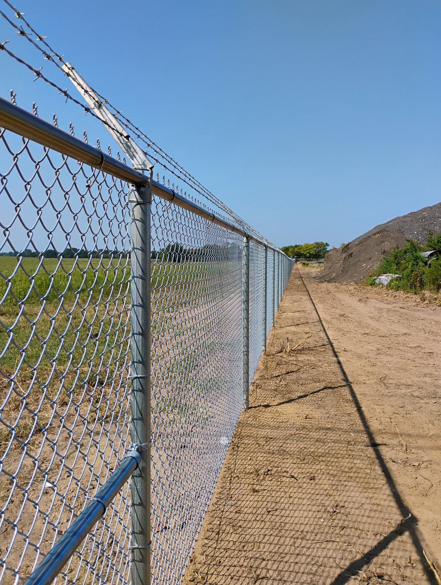 Chain-link fence beside a dirt path under a blue sky, with hillside terrain in the distance.