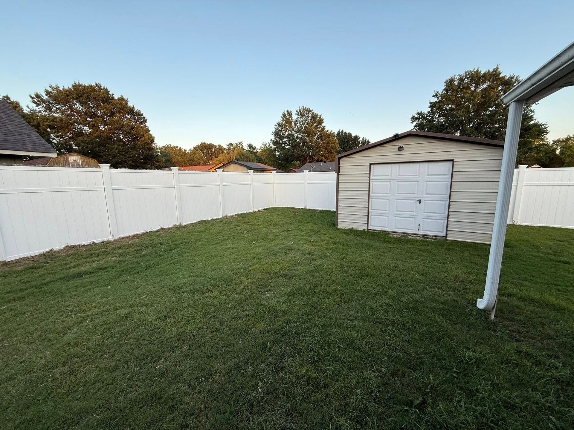 Backyard with white fence, green lawn, and a small gray shed under a blue sky